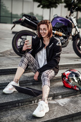 Young beautiful and stylish smiling girl in glasses sits on the steps near her purple motorcycle and looks at the screen of her mobile phone.