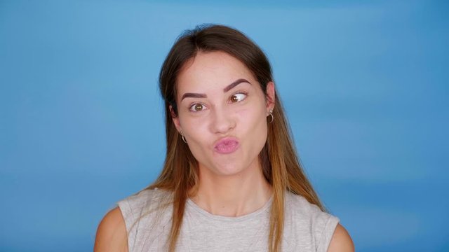 Beautiful funny woman in gray t-shirt squints and smiles on blue background with copy space. 4K slow motion footage