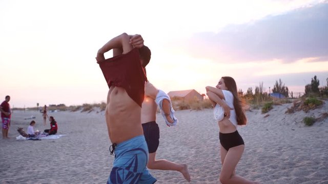 Group of happy friends undress running into the sea water and throw shirts on the beach. Cheerful people having fun on shore near ocean. Young men and women go to swim and bathe. 120 fps slow motion.