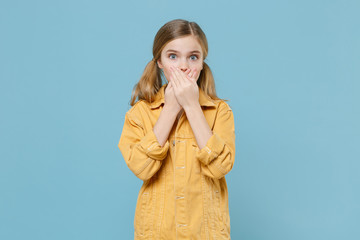 Shocked little blonde kid girl 12-13 years old in yellow jacket isolated on pastel blue background children studio portrait. Childhood lifestyle concept. Mock up copy space. Covering mouth with hands.