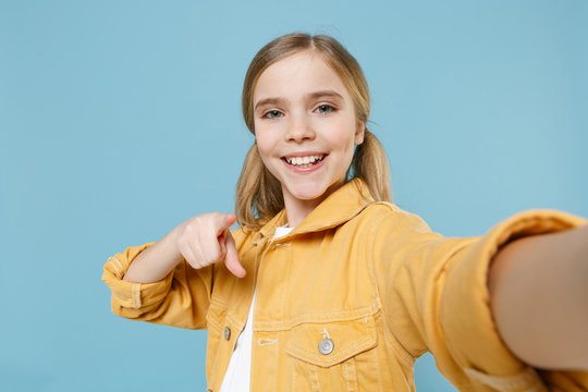 Close up of smiling little blonde kid girl 12-13 years old in yellow jacket isolated on blue background. Childhood lifestyle concept. Doing selfie shot on mobile phone pointing index finger on camera.