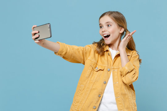 Excited little blonde kid girl 12-13 years old in yellow jacket isolated on pastel blue background. Childhood lifestyle concept. Mock up copy space. Doing selfie shot on mobile phone, spreading hands.