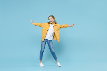 Full length portrait of cheerful little blonde kid girl 12-13 years old in yellow jacket isolated on blue background studio portrait. Childhood lifestyle concept. Mock up copy space. Spreading hands.
