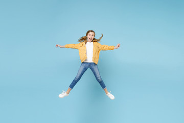 Fototapeta na wymiar Full length portrait of excited little blonde kid girl 12-13 years old in yellow jacket isolated on blue background studio. Childhood lifestyle concept. Mock up copy space. Jumping, spreading hands.