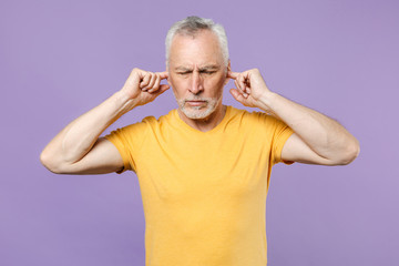 Displeased elderly gray-haired mustache bearded man in yellow t-shirt posing isolated on pastel violet wall background. People lifestyle concept. Covering ears with fingers, keeping eyes closed.