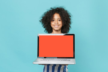 Smiling little african american kid girl 12-13 years old in striped clothes isolated on blue background. Childhood lifestyle concept. Mock up copy space. Hold laptop computer with blank empty screen.