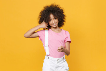 Obraz na płótnie Canvas Smiling little african american kid girl 12-13 years old in pink t-shirt isolated on yellow background. Childhood lifestyle concept. Doing phone gesture like says call me back, point finger on camera.