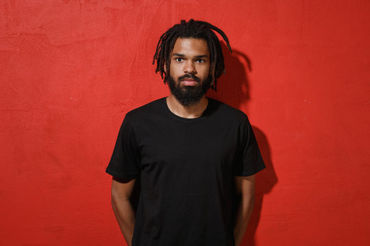 Handsome attractive young african american man guy with dreadlocks 20s wearing black casual t-shirt posing looking camera isolated on bright red color wall background studio portrait.