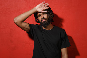 Exhausted tired young african american man guy with dreadlocks 20s wearing black casual t-shirt posing put hand on head looking camera isolated on bright red color wall background studio portrait.