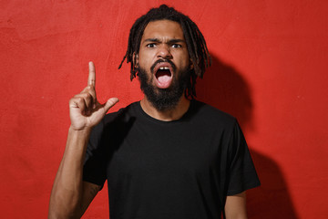 Irritated young african american man guy with dreadlocks 20s in black casual t-shirt posing screaming swearing pointing index finger up looking camera isolated on red color background studio portrait.