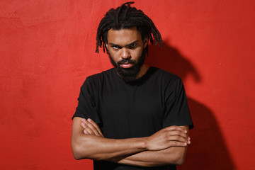 Displeased offended concerned young african american man guy with dreadlocks 20s wearing casual black t-shirt posing holding hands crossed isolated on bright red color wall background studio portrait.