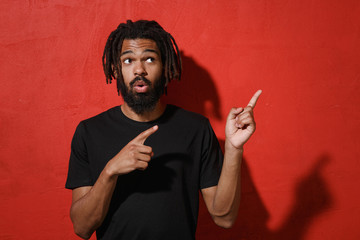Shocked amazed young african american man with dreadlocks 20s in black casual t-shirt posing point index fingers aside up on mock up copy space isolated on bright red color background studio portrait.