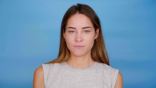 Positive woman in gray t-shirt smiles, raises hands and shows middle finger on blue background with copy space. 4K slow motion footage