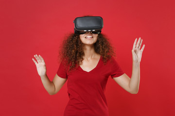 Young african american girl in casual t-shirt posing isolated on red background. People lifestyle concept. Watching in headset, touch something like push on button point at floating virtual screen.