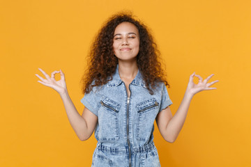 Smiling young african american girl in denim clothes isolated on yellow background. People lifestyle concept. Mock up copy space. Hold hands in yoga gesture, relaxing meditating, trying to calm down.