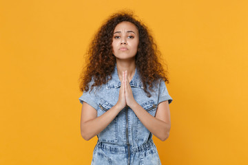 Obraz premium Pleading young african american woman girl in casual denim clothes isolated on yellow background studio portrait. People emotions lifestyle concept. Mock up copy space. Holding hands folded in prayer.