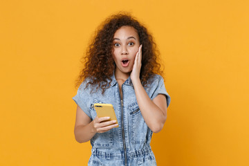 Shocked young african american girl in casual denim clothes isolated on yellow background studio portrait. People lifestyle concept. Mock up copy space. Using mobile cell phone, put hand on cheek.