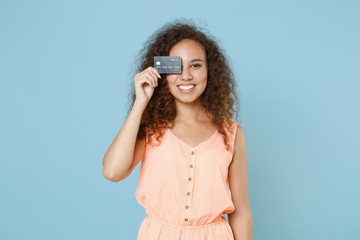 Smiling young african american woman girl in pastel summer clothes posing isolated on blue wall background studio. People lifestyle concept. Mock up copy space. Covering eye with credit bank card.