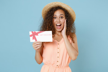 Shocked young african american girl in pastel summer clothes hat posing isolated on blue wall background studio. People lifestyle concept. Mock up copy space. Hold gift certificate put hand on cheek.