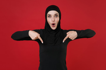 Shocked young arabian muslim woman in hijab black clothes posing isolated on red wall background studio portrait. People religious lifestyle concept. Mock up copy space. Pointing index fingers down.