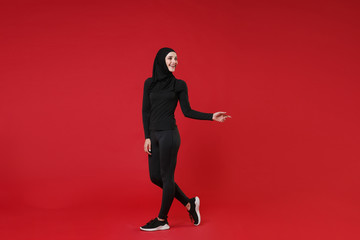 Fototapeta na wymiar Full length portrait of smiling young arabian muslim woman in hijab black clothes isolated on red wall background. People religious lifestyle concept. Mock up copy space. Pointing index finger aside.
