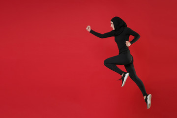 Fototapeta na wymiar Full length portrait side view of young arabian muslim woman in hijab black clothes posing isolated on red wall background studio portrait. People religious lifestyle concept. Jumping like running.