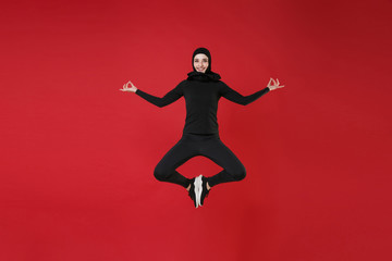 Fototapeta na wymiar Full length portrait of smiling young arabian muslim woman in hijab black clothes posing isolated on red background studio. People religious lifestyle concept. Jumping, hold hands in yoga gesture.