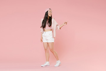 Full length portrait of smiling young asian girl in casual clothes, cap isolated on pastel pink background studio portrait. People lifestyle concept. Mock up copy space. Pointing index finger aside.