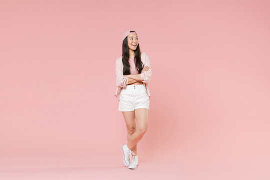 Full length portrait of smiling young asian girl in casual clothes, cap isolated on pastel pink background studio. People lifestyle concept. Mock up copy space. Holding hands crossed, looking aside.