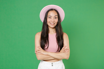 Fototapeta premium Smiling young asian woman girl in casual pink clothes hat posing isolated on green background studio portrait. People sincere emotions lifestyle concept. Mock up copy space. Holding hands crossed.