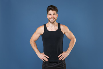 Smiling young bearded fitness sporty guy sportsman in black undershirt isolated on blue wall background studio portrait. Workout sport motivation lifestyle concept. Standing with arms akimbo on waist.