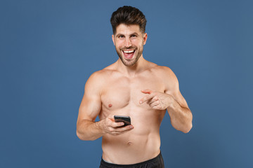Cheerful young bearded fitness sporty strong guy bare-chested muscular sportsman isolated on blue background. Workout sport motivation lifestyle concept. Pointing index finger on mobile cell phone.