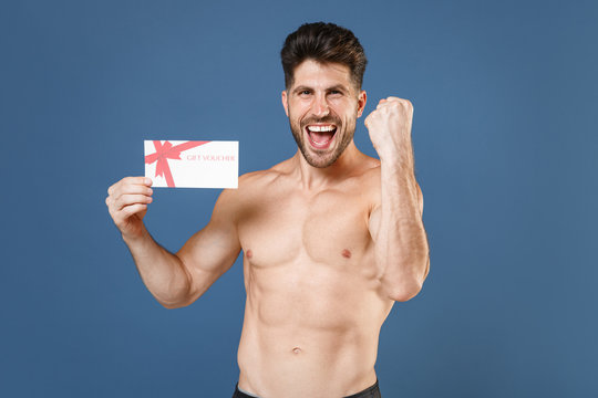 Happy young bearded fitness sporty strong guy bare-chested muscular sportsman isolated on blue wall background. Workout sport motivation lifestyle concept. Hold gift certificate, doing winner gesture.