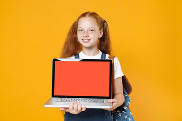 Naklejka premium Young redhead school girl 12-13 years old in white t-shirt blue denim uniform backpack hold laptop pc computer isolated on yellow background children studio portrait Kids education lifestyle concept