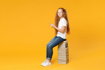 Full length redhead young girl 12-13 years old in white tshirt sit on big stack school book hold using mobile cell phone isolated on yellow background children studio portrait Kids education concept