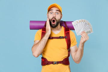 Shocked traveler young man in t-shirt cap backpack isolated on blue background. Tourist traveling on weekend getaway. Tourism discovering hiking concept. Hold fan of cash money in dollar banknotes.