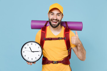 Cheerful young traveler man in yellow casual t-shirt cap backpack isolated on blue background. Tourist traveling on weekend getaway. Tourism discovering hiking concept. Hold clock showing thumb up.