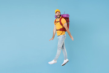 Full length portrait Excited young traveler man in cap with backpack isolated on blue background. Tourist traveling on weekend getaway. Tourism discovering hiking concept. Jump spreading hands legs.