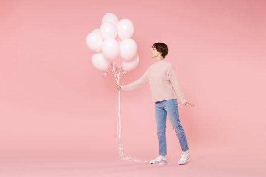 Full length portrait side view of smiling young woman in casual sweater isolated on pastel pink background. Birthday holiday party people emotions concept. Celebrating hold air balloons looking aside.