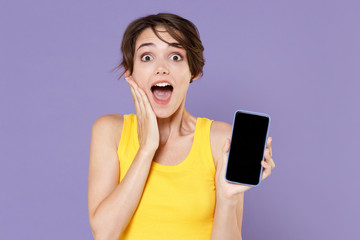Naklejka premium Shocked young woman in yellow casual tank top isolated on violet background studio portrait. People lifestyle concept. Mock up copy space. Hold mobile phone with blank empty screen put hand on cheek.