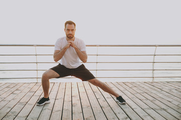 Full length portrait of handsome young bearded athletic man guy 20s in white t-shirt posing training doing stretching lunge exercising for legs looking camera at sunrise over the sea outdoors. - obrazy, fototapety, plakaty