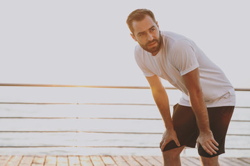 Portrait of tired handsome attractive young bearded athletic man guy 20s in white t-shirt posing training standing resting hold hands on knees looking aside at sunrise over the sea outdoors.