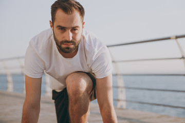 Portrait of handsome young bearded athletic man guy 20s in white t-shirt black shorts listening music with air pods training standing at low start before running at sunrise over the sea outdoors.