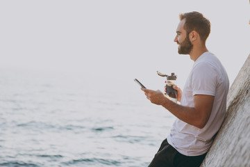 Portrait smiling young bearded fitness athletic man guy 20s in casual white t-shirt posing training resting using mobile cell phone hold bottle of water looking aside at sunrise over the sea outdoors.
