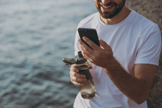 Cropped image portrait smiling young bearded fitness athletic man guy 20s in casual white t-shirt resting using mobile cell phone hold bottle of water looking aside at sunrise over the sea outdoors.