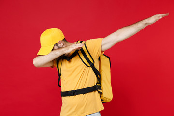 Delivery man 20s in yellow cap t-shirt thermal bag backpack DAB dance gesture isolated on red background studio Guy male employee work as courier Service quarantine coronavirus covid-19 virus concept - obrazy, fototapety, plakaty