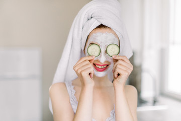 Funny young Caucasian girl with clay mask for face skin and cucumbers on eyes, posing at light home kitchen and having fun, enjoying spa beauty procedures at home
