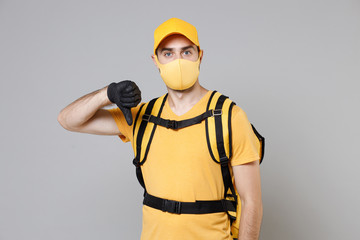 Delivery employee man guy in yellow cap t-shirt uniform face mask gloves thermal food bag backpack work courier service during quarantine coronavirus covid-19 virus isolated on gray background studio.