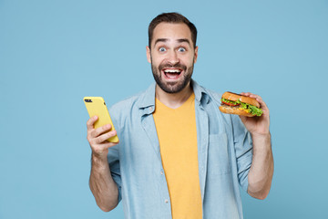 Surprised young bearded man guy 20s wearing casual clothes posing holding in hands american classic fast food burger using mobile cell phone isolated on pastel blue color background studio portrait.