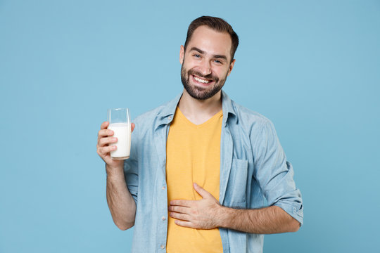 Smiling Young Bearded Man Guy 20s Wearing Casual Clothes Posing Holding In Hand Glass Of Milk Put Hand On Stomach Looking Camera Isolated On Pastel Blue Color Wall Background Studio Portrait.